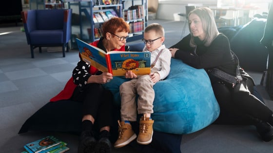 Volontären och läsfaddern Lena Olsson och Eman Begic sitter på kuddar och läser en bok. Bredvid sitter Emans mamma Alma.