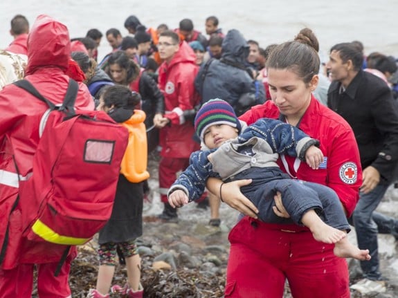 En Röda Korset-volontär som bär ett barn vid strandkanten. I bakgrunden syns en båt som precis kommit i land full med människor.